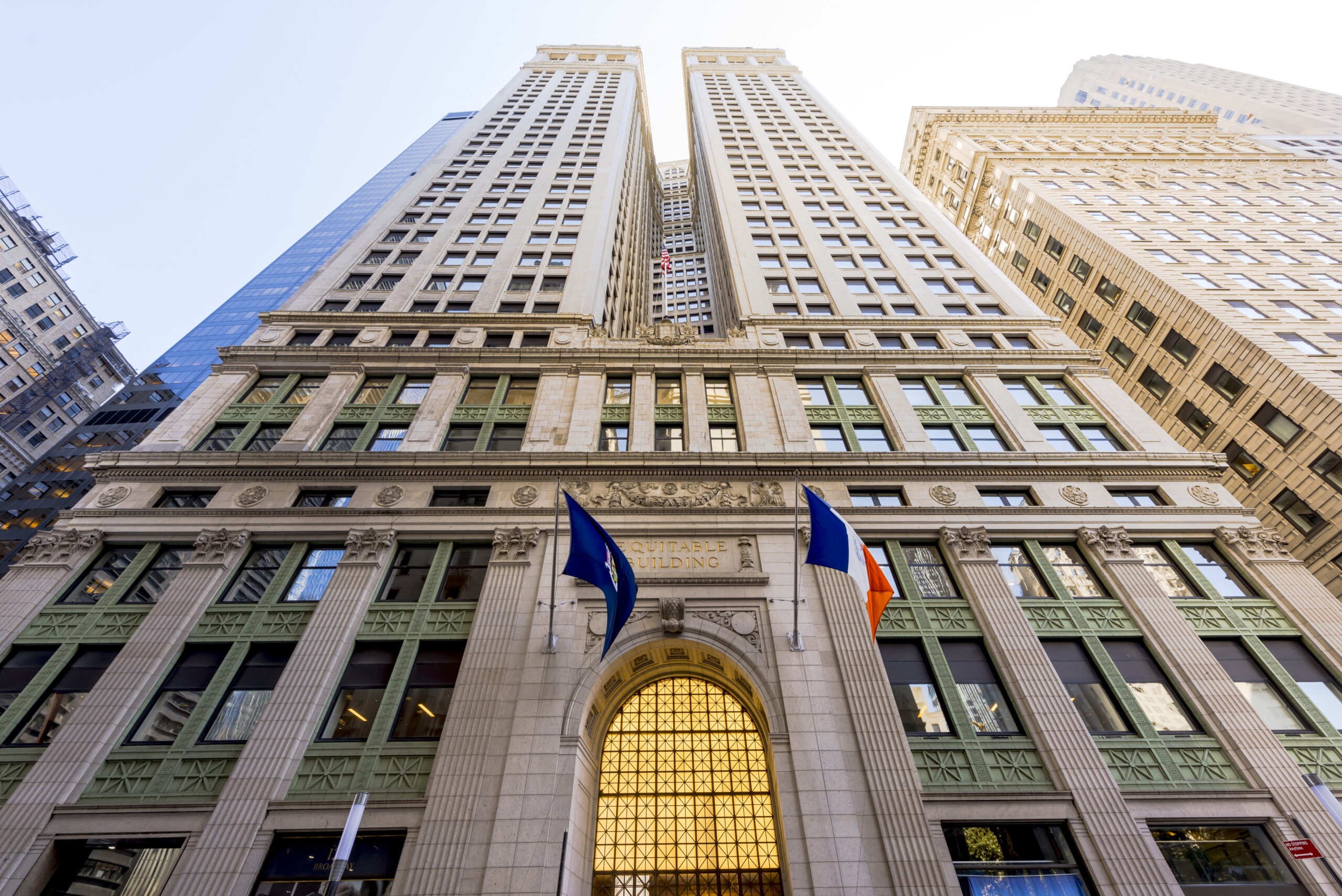 Low-angle view of a tall, ornate office building in Canada, with arched entrance, green window frames, decorative stonework, and three flags above the doorway; ideal for lawyers specializing in immigration as neighboring skyscrapers rise on both sides.