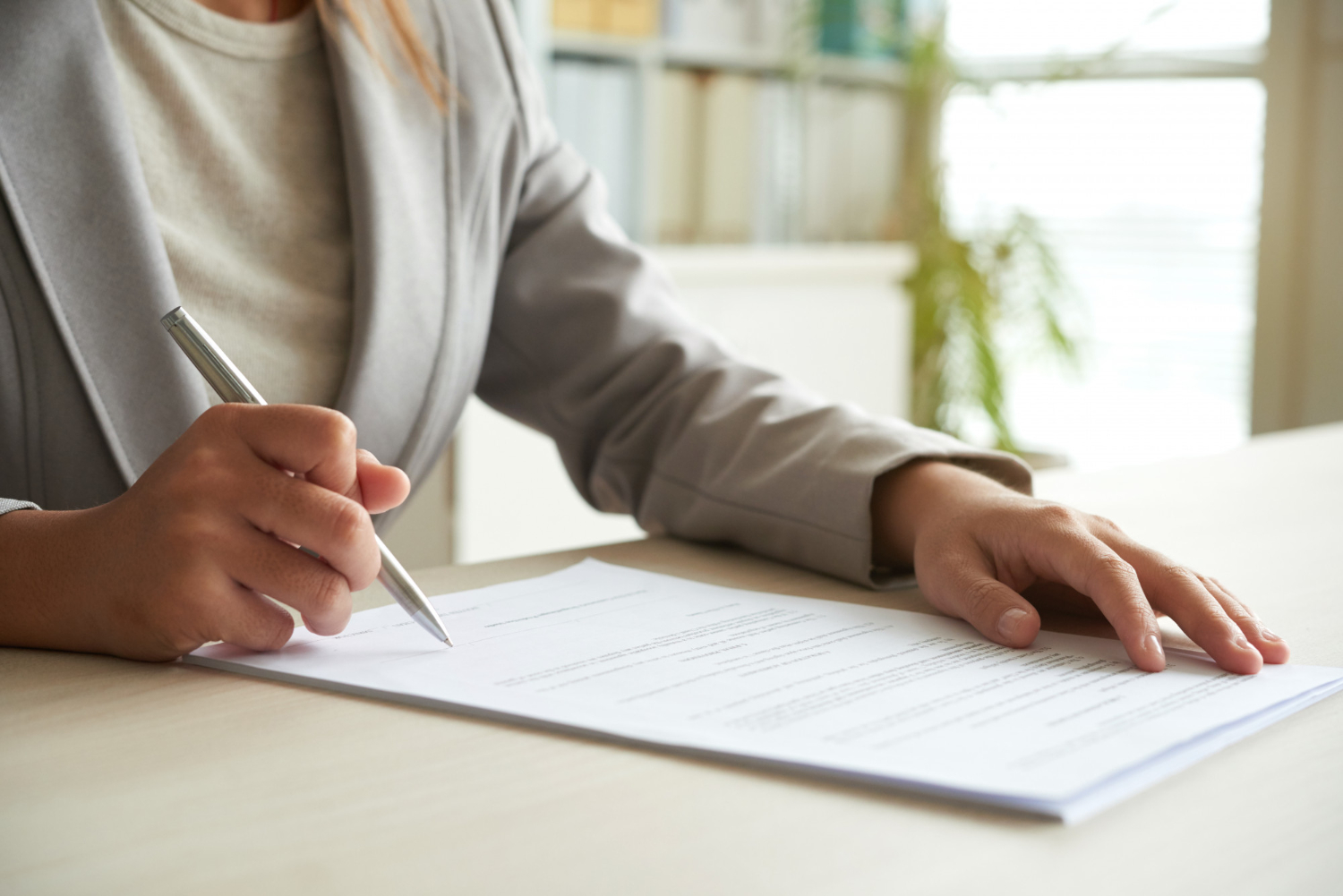 Person in a light gray blazer sitting at a desk, holding a pen and signing an immigration document on a clipboard—an image often associated with lawyers handling important paperwork.